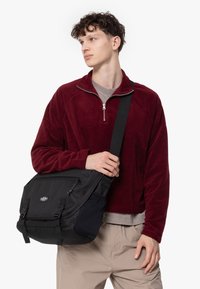 Borsa a tracolla nera con lati imbottiti, tracolla regolabile e cerniere. Indossata da una persona in un pullover di pile bordeaux e pantaloni beige.