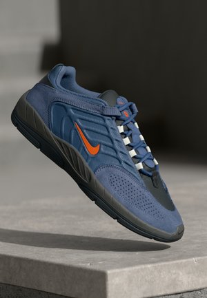 Zapatilla Nike azul y negra con el logotipo naranja y cordones azules flotando sobre una superficie de hormigón gris claro.
