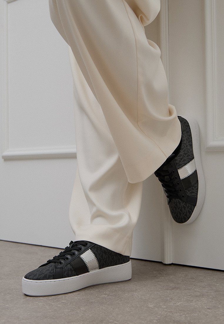 Zapatillas negras y grises con una parte superior texturizada, acentos blancos y una suela gruesa blanca. Llevadas con pantalones color crema frente a un fondo neutro.