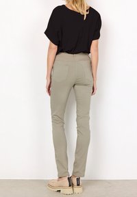 Femme debout face à un mur portant un haut noir à manches courtes, un pantalon slim kaki et des chaussures beiges à semelle épaisse avec des languettes noires au talon.