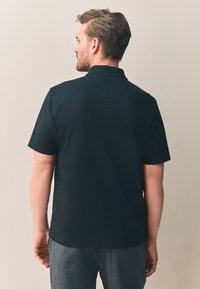 Donker poloshirt getoond op een mannelijk model, met korte mouwen, een kraag en een gladde, effen textuur met een licht aansluitende pasvorm.