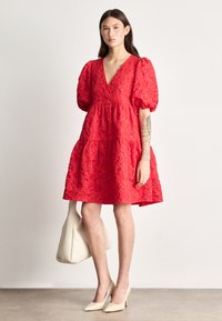 Raudona gėlėmis siuvinėta suknelė su giliu V formos dekolte, pūsto tipo rankovėmis ir sluoksniuota suknele; derinama su balta rankine ir kūno spalvos aukštakulniais.