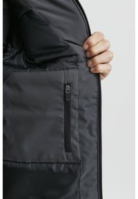 Svart vadderad jacka inuti med en ficka med dragkedja, slät textur och minimalt med sömmar. Har en mjuk fodring och dold dragkedja.