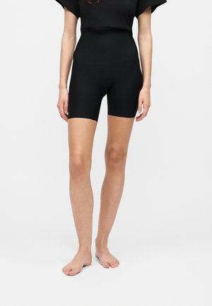 Femme portant un short de biker noir moulant et un haut noir assorti, debout pieds nus sur un fond blanc uni.