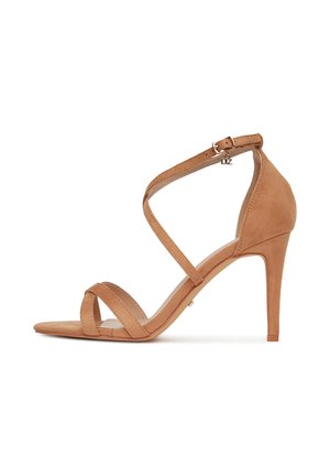 Sandalo con tacco alto in camoscio color cuoio con punta aperta, caratterizzato da cinturini incrociati e chiusura con fibbia alla caviglia. Texture liscia e tacco affusolato.