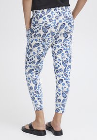 Witte broek met een blauw paisleypatroon, smal toelopende fit en twee zijzakken. Gecombineerd met zwarte sandalen.