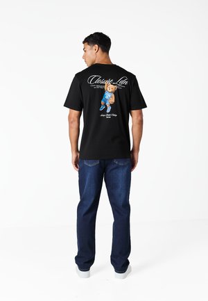 Άνδρας που φοράει μαύρο μπλουζάκι με εκτύπωση αρκουδάκι ντυμένο με μπλε αθλητική στολή στο πίσω μέρος, συνδυασμένο με μπλε τζιν και λευκά παπούτσια, κοιτάζοντας προς τα πίσω.