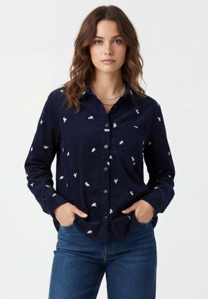 Femme aux cheveux bruns ondulés portant une chemise bleu marine boutonnée avec des motifs floraux blancs et un jean bleu, les mains dans les poches, sur un fond uni.