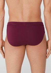 Burgundské pánské slipy s plochým pasem, hladkým povrchem a těsným střihem, které ukazují bezešvý design a minimální pokrytí nohavic.