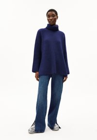 Blauer gerippter Rollkragenpullover mit lockerer Passform, kombiniert mit weit geschnittenen, hellblauen Denim-Jeans mit seitlichen Schlitzen und silbernen Sneakers.