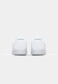 Tênis brancos de couro com textura lisa, apresentando um logo verde "LACOSTE" no calcanhar e uma sola plana de borracha cinza.