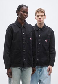Schwarze Jacken mit Knopfleiste aus robustem Stoff, mit aufgesetzten Taschen, silbernen Knöpfen und einem kleinen mehrfarbigen Logo auf der linken Brust.