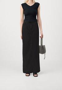 Kvinna i svart figursydd V-ringad ärmlös topp, lång svart kjol, svarta öppna skor, som håller en liten pärlhandväska framför vit bakgrund.