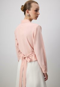 Light pink long-sleeve blouse with a collar and back tie detail, made of smooth fabric. Buttoned cuff accents. Simple design.