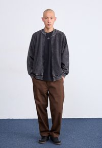 Casaco bomber cinza com listas laterais, t-shirt preta por baixo, calças largas castanhas e sapatilhas pretas. Design simples contra um fundo claro.