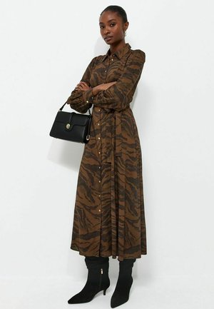 Mujer con un vestido largo marrón con estampado y botones, botas negras hasta el tobillo, sosteniendo un bolso negro sobre un fondo blanco liso.