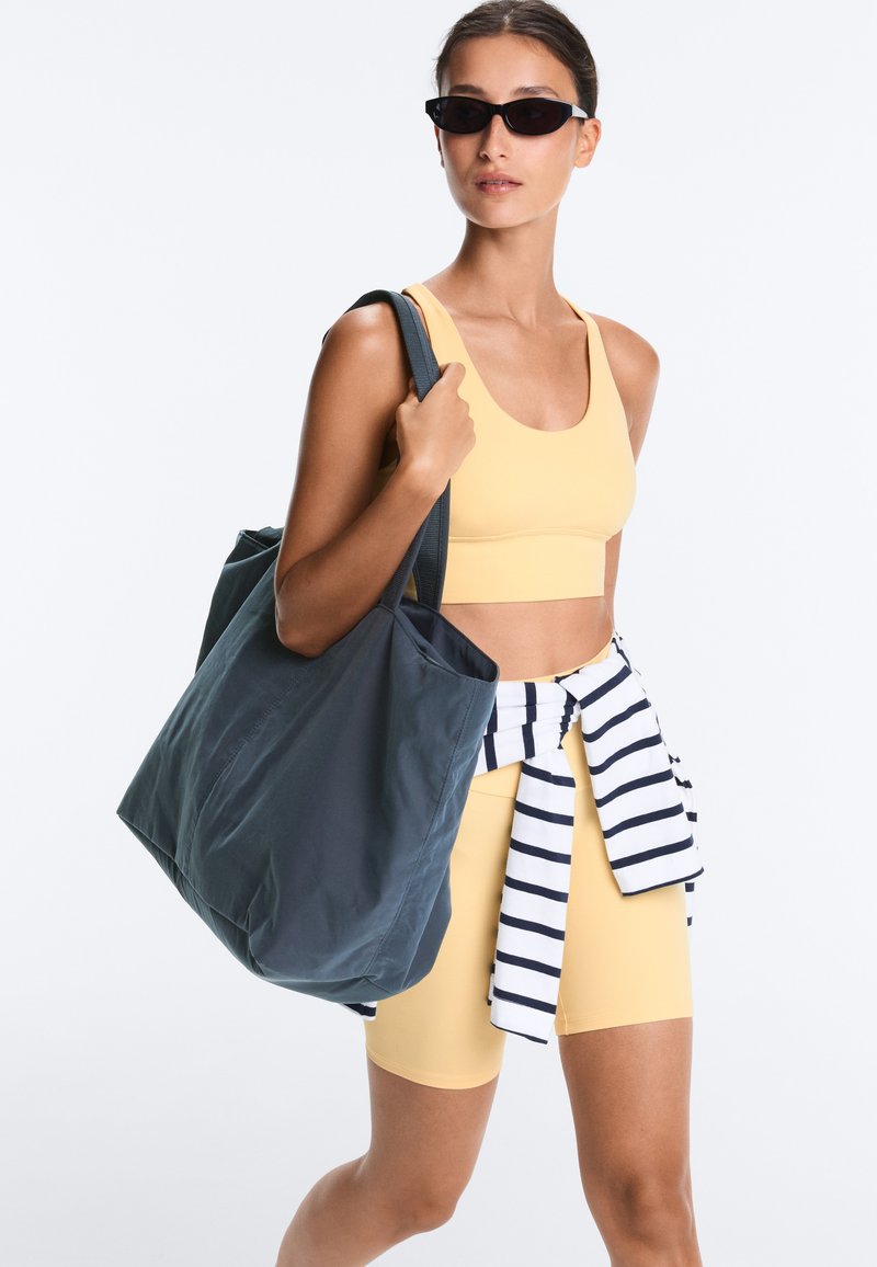Vrouw met zonnebril, gele sportbeha en korte broek, gestreepte trui gestrikt om de taille, draagt een grote donkere tas aan de schouder.
