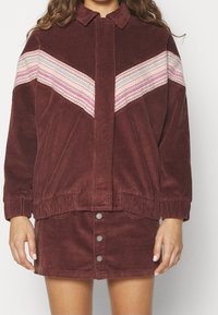 Vrouw in een bordeaux corduroy jack en bijpassende rok, het jack heeft roze, witte en grijze chevronstrepen over de borst en op de mouwen.