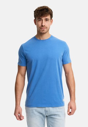 Junger Mann mit braunen lockigen Haaren, der ein schlichtes blaues T-Shirt mit kurzen Ärmeln und hellblaue Jeans trägt, vor einem weißen Hintergrund stehend.