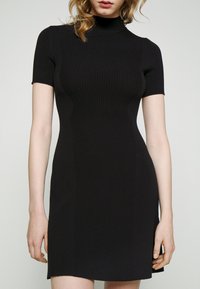 Femme portant une robe noire moulante à manches courtes côtelée avec un col haut, debout devant un fond clair uni.