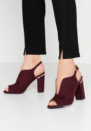 Fødder iført bordeaux ruskind åbne slingback-højhælede sko med blokhæl, kombineret med sorte korte bukser med små sideslids.