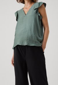 Ljusgrön V-ringad topp med volangärmar, som har en avslappnad passform och texturerad tyg. Matchad med svarta, vida byxor.
