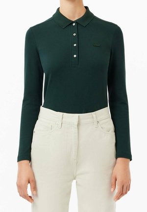 Donkergroene polo met lange mouwen, vier witte knopen en een logo op de borst, gedragen ingestopt in een hoog getailleerde off-white broek.