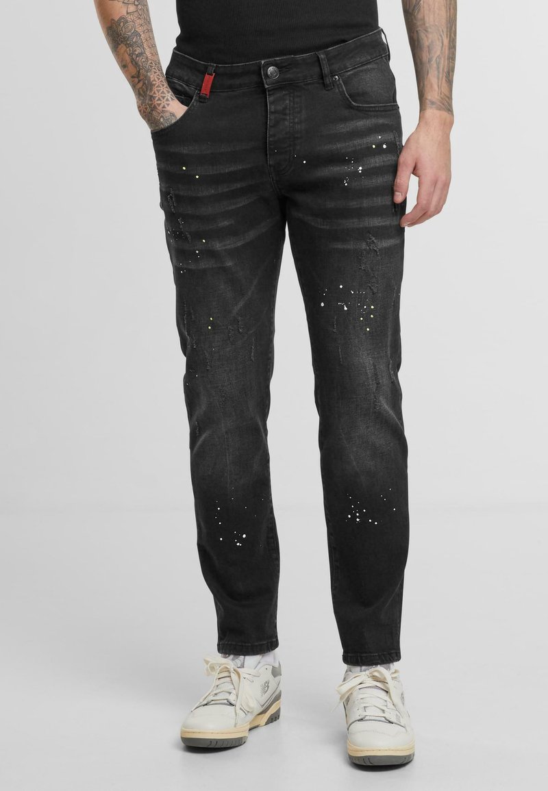 Jeans de mezclilla negra con corte slim, que presentan un desgaste sutil, detalles desgastados y salpicaduras de pintura blanca dispersas.