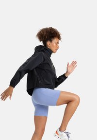 Black lightweight jacket with a hood, elastic cuffs, and a yellow zipper, paired with light blue cycling shorts. Model in a running pose.