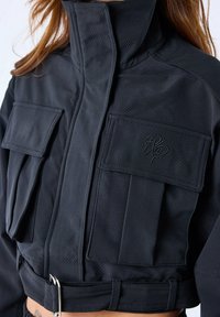 Zwarte cropped jacket met een hoge kraag, twee voorkruiszakken, een textuurpatroon en een gespdetail in de taille.