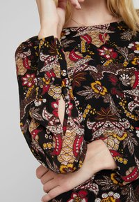 Robe noire à manches longues avec des motifs floraux et paisley en rouge, jaune et blanc. Équipée de poignets boutonnés et d'un tissu texturé.