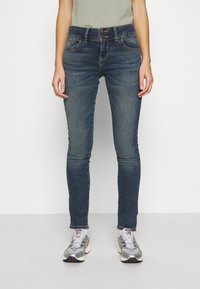 Blaue Jeans im Skinny-Schnitt mit hoher Taille, zwei vorderen Taschen und einer leichten Verwässerung. Getragen mit grauen Sportschuhen mit reflektierenden Akzenten.