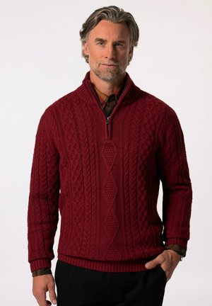 Maglione lavorato a maglia bordeaux con design a trecce, colletto mezza zip, polsini e orlo a coste, indossato sopra una camicia scozzese. Dettaglio testurizzato lungo tutto il capo.