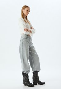 Valkoinen pitkähiha blouse, jossa on röyhelöillä varustetut hihansuut, yhdistettynä vaaleanharmaisiin leveisiin farkkuihin. Mustat cowboy-kengät, joissa on kohokuviointi ja neliön muotoinen kärki.