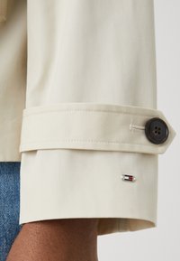 Beżowy mankiet płaszcza z czarnym guzikiem i małą przypinką z logo Tommy Hilfiger, noszony na ciemnoskórym nadgarstku oraz niebieskich dżinsach.