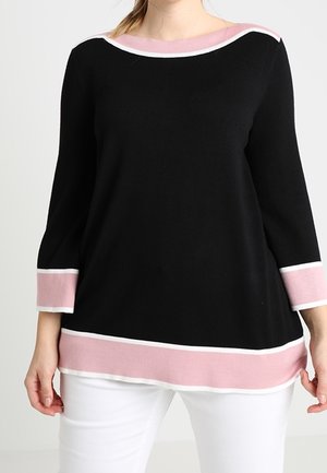 Kvinde iført en sort sweater med lyserøde og hvide kanter på manchetter, nederkant og halsudskæring, kombineret med hvide bukser mod en ensfarvet baggrund.