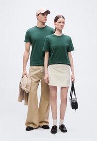 Άνδρας και γυναίκα φορούν τα ίδια πράσινα μπλουζάκια· ο άνδρας κρατάει μπεζ σακάκι, η γυναίκα έχει μαύρη τσάντα χειρός, στέκονται μπροστά σε απλό φόντο.