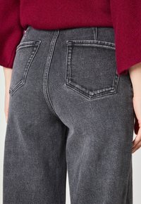 Jean taille haute, coupe droite en denim gris foncé, avec deux poches arrière ornées de coutures et un tissu lisse et texturé.