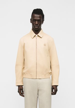 Mand iført en beige jakke med lynlås og krave samt beige bukser, stående foran en ensartet lys baggrund.