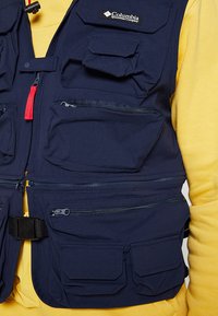 Gilet blu navy realizzato in tessuto resistente, dotato di molteplici tasche frontali, un compartimento con zip e una cintura nera con fibbia in plastica.