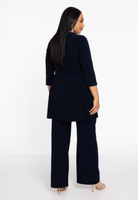 Marineblauwe tweedelige outfit met een lange tuniek top en wijde broekspijpen van zachte stof. Beschikt over 3/4 mouwen en een gladde textuur, gecombineerd met huidskleurige hakken.