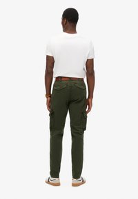 Hombre con camiseta blanca de manga corta, pantalones cargo verde oliva con bolsillos laterales, cinturón marrón y zapatillas blancas, de pie de espaldas sobre un fondo liso.