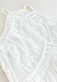 Robe blanche sans manches avec broderie œillets géométrique et bordure perlée texturée sur tissu léger.