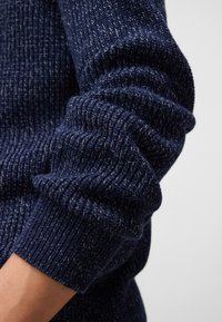 Marineblauer Strickpullover mit gerippter Textur und geknöpften Ärmeln, der subtile Abweichungen in der Fadenfarbe aufweist, um zusätzliche Tiefe zu verleihen.