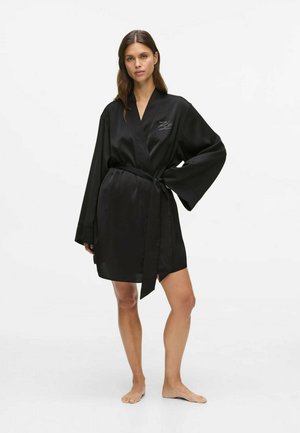 Le modèle pose pieds nus dans un peignoir en satin noir ceinturé, à larges manches et détail brodé sur la poitrine, sur un fond blanc de studio.