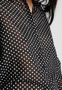 Black, sheer blouse with white polka dots, buttoned design, and a collar. Notable texture with a lightweight, draping appearance.