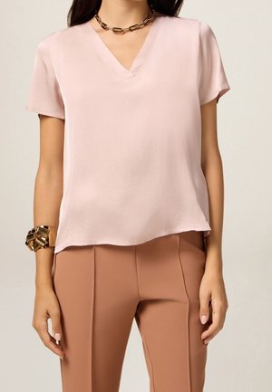 Frau trägt eine hellrosa V-Ausschnitt-Bluse mit kurzen Ärmeln, rostfarbene Hose, ein massives goldenes Armband und eine goldene Kettenhalskette.