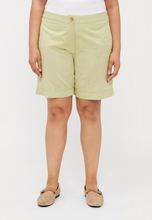 Pantaloni corti verde chiaro in tessuto testurizzato, con un unico bottone frontale e orlo arrotolato, abbinati a scarpe beige.