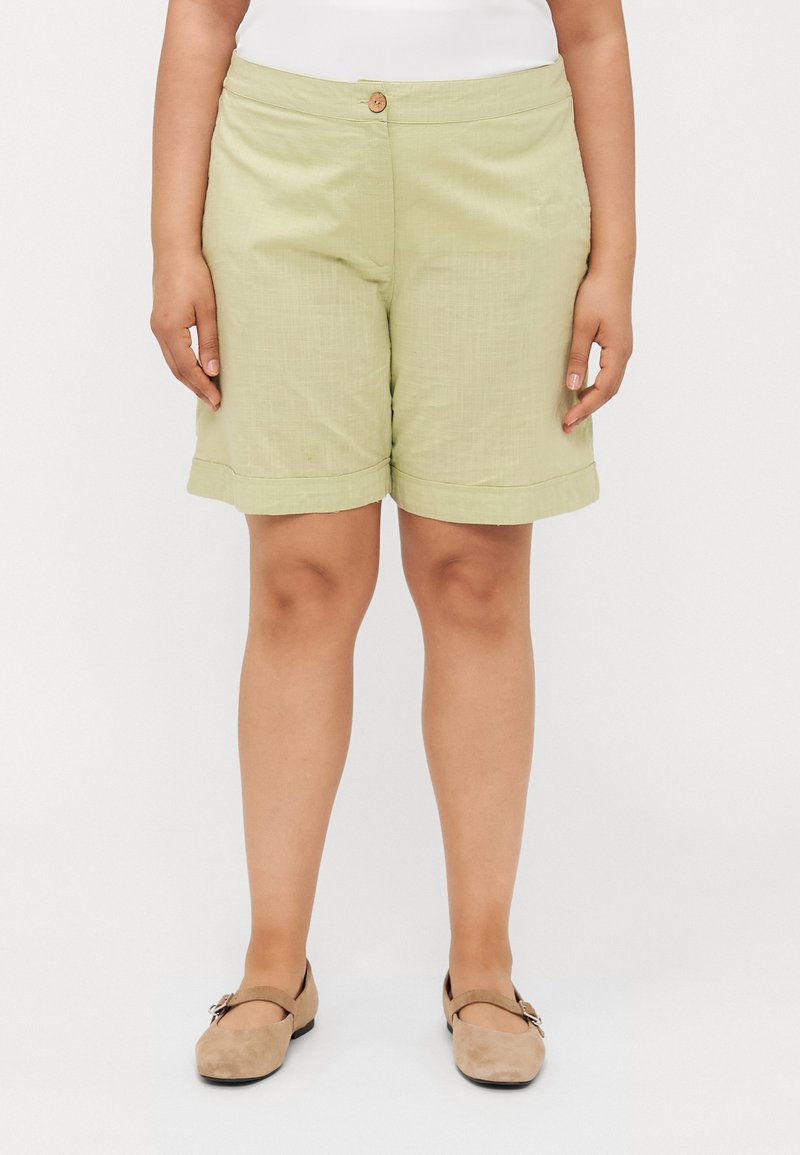 Shorts verde claro elaborados con tejido texturizado, que cuentan con un único botón delantero y dobladillo vuelto, combinados con zapatos beiges.