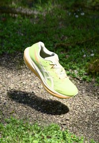 Light green running shoe with a textured upper, white accents, and a light orange sole, suspended above gravel and grass.
